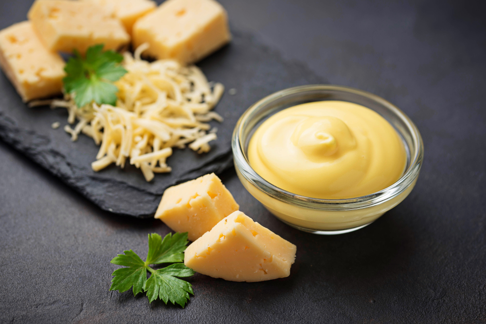 Un recipient de sos, în care se află sos de brânză, alături de bucăți de brânză și brânză răzuită.