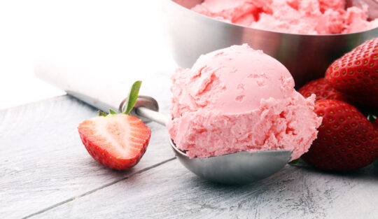 Înghețată de căpșuni pe o linguriță din inox, cu căpșuni tăiate alături