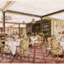 Sala de mese a vasului Titanic, cu scaune cu tapițerie scumpă, aurie și mese rotunde, cu acoperite cu fețe de masă de un alb imaculat.