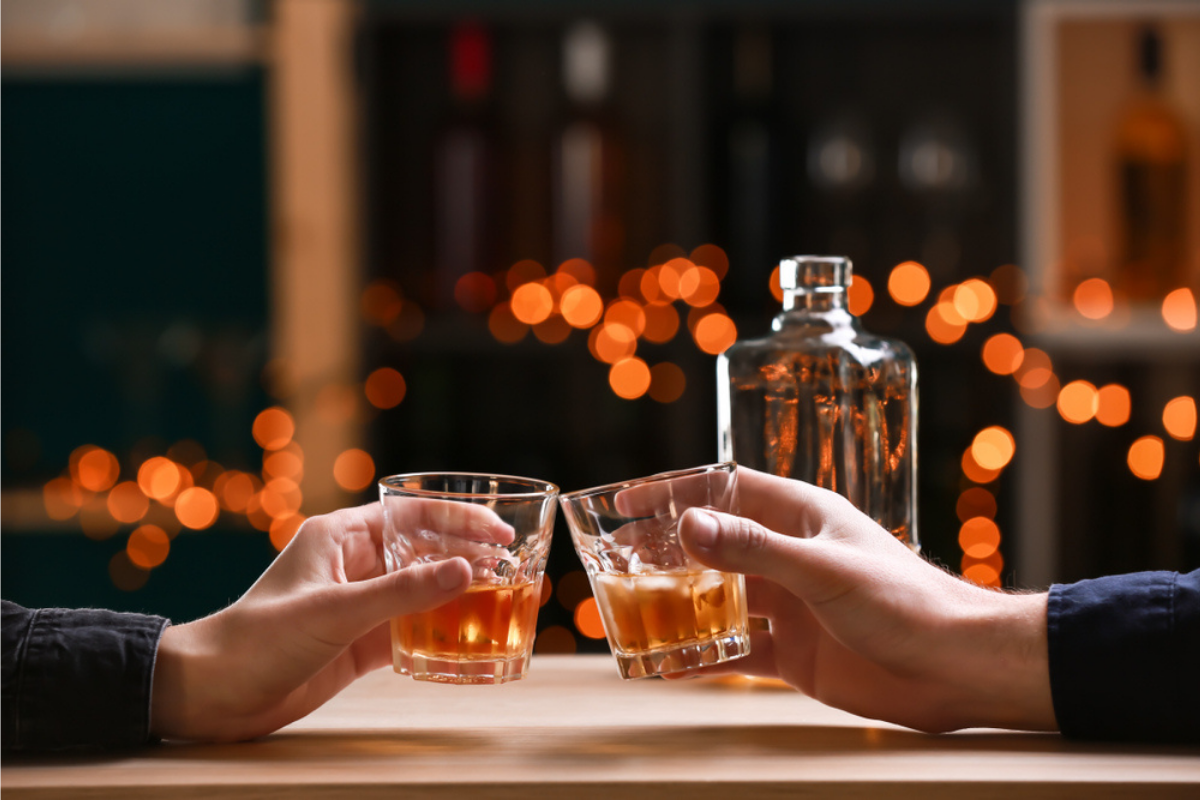 sticlă de alcool deschisă în fața a două persoane ciocnind pahare