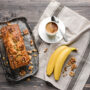 Banana bread, într-o tavă de chec, cu o cafea și o banană alături, pe un prosop de bucătărie