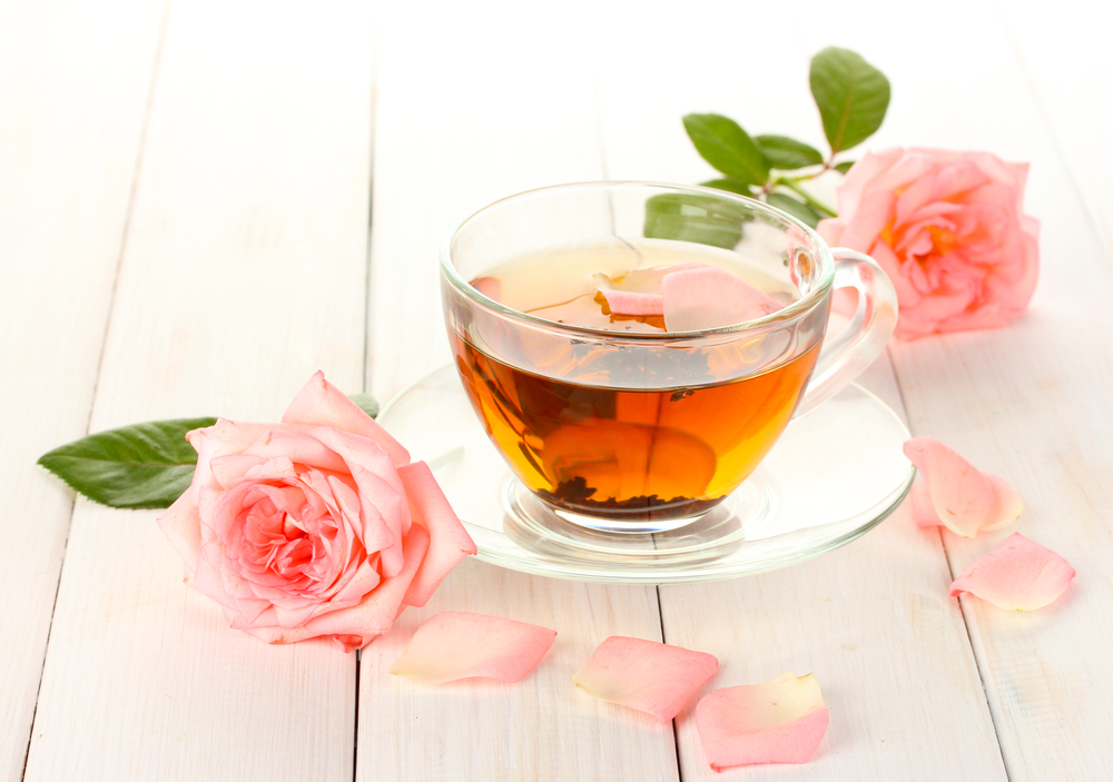 O ceașcă cu ceai verde și trandafiri, așezată pe o masă alături de doi trandafiri de culoare roz.