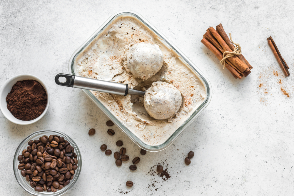 Înghețată cu aromă de cafea, așezață într-un recipient dreptunghiular, alături de boabe de cafea, scorțișoară și cafea râjnită.