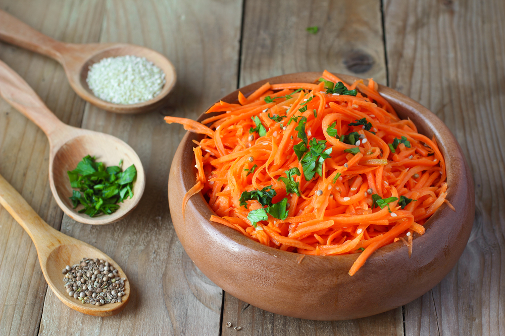 Salata de morcovi pusă într-un bol de lemn alături de trei linguri cu condimente