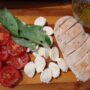 Piept de pui crestat pe un tocător, alături de felii de roșii, felii de mozzarella, frunze de busuioc și un bol cu ulei de măsline și oregano