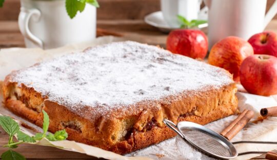 Prăjitură cu mere, decorată cu zahăr, pe o hârtie de copt, alături de mere, ceașcă albă, sită pentru zahăr și un ghiveci cu frunze de mentă