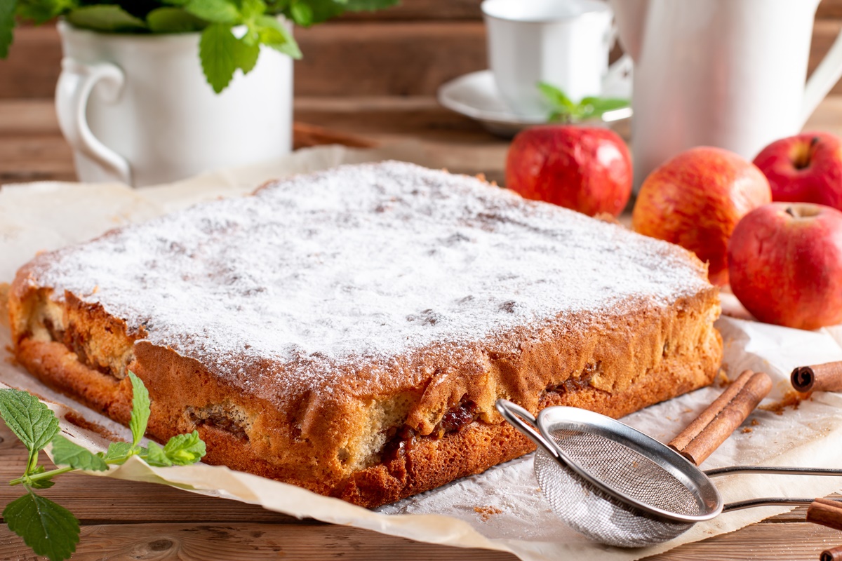 Prăjitură cu mere, decorată cu zahăr, pe o hârtie de copt, alături de mere, ceașcă albă, sită pentru zahăr și un ghiveci cu frunze de mentă