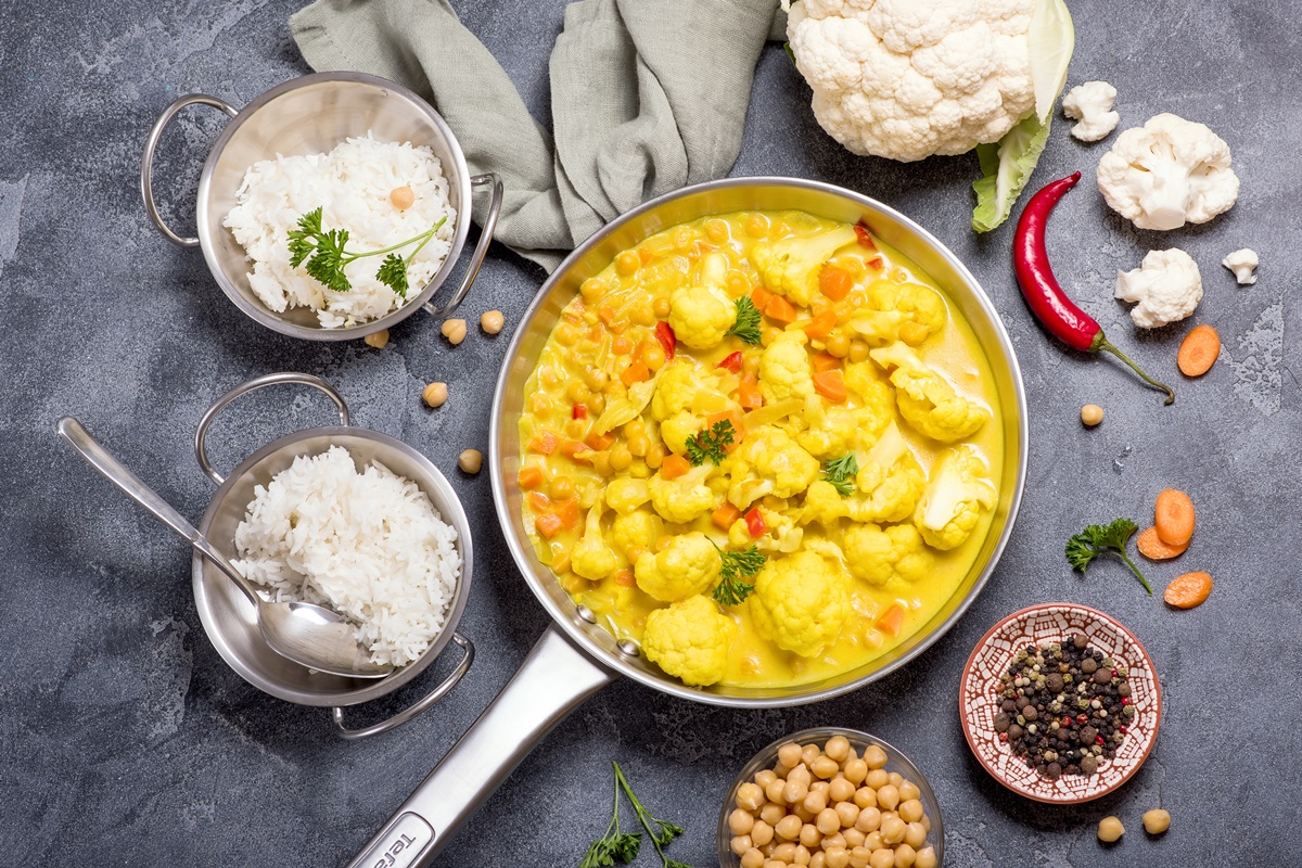 Curry de năut și conopidă într-o tigaie, alături de două boluri cu orez , un bol cu năut, unul cu piper și o conopidă