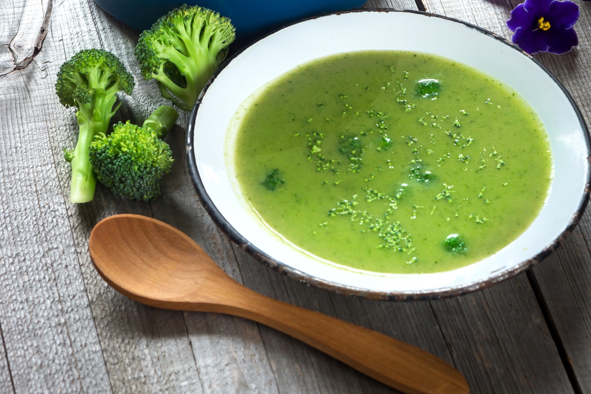 Castron metalic cu supă cremă de broccoli alături de bucăți de broccoli proaspăt și lingură de lemn