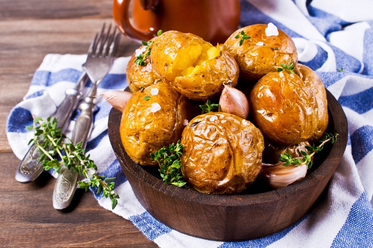 Bol cu cartofi copți în coajă, presărați cu usturoi și cimbru verde, pe un ștergar alb cu albastru, alături de tacâmuri de servire