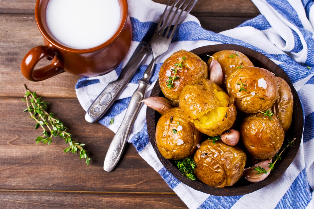 Cartofi întregi la cuptor, cu cimbru și usturoi, așezați într-un bol pe un ștergar alb cu albastru, alături de tacâmuri și o cană cu smântână