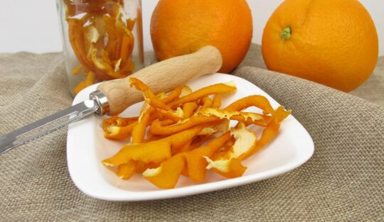 Coji de portocale în borcan și în farfurie, alături de dispozitiv de curățare și două portocale