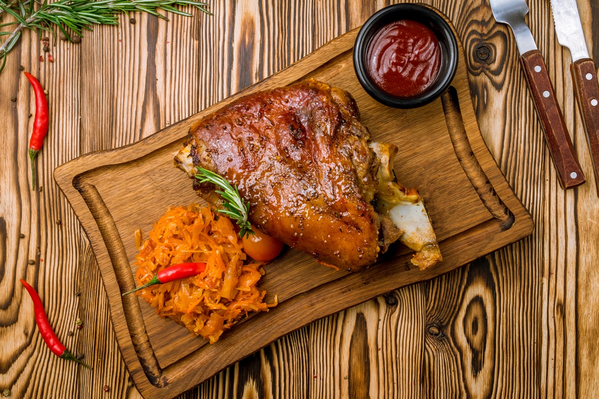 Porție de varză murată cu ciolan afumat pe o tavă de lemn, alături de ardei iuți și un bol negru cu sos de roșii