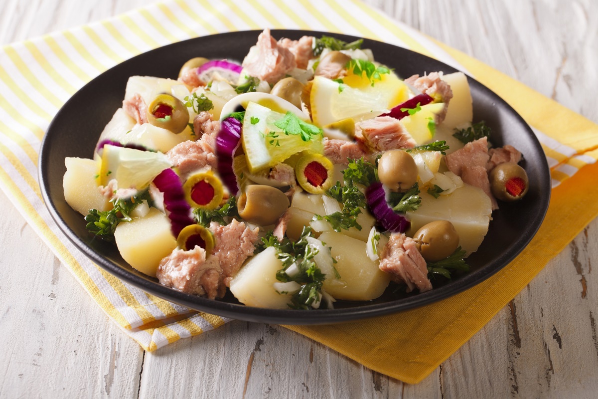 Salată de cartofi cu ton și măsline umplute cu ardei în farfurie neagră așezată pe două ștergare
