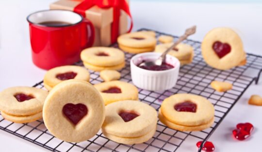 Biscuiți cu forme de inimioară și jeleu de zmeură pe un grătar, alături de o cană roșie, o cutie de cadouri și un bol cu dulceață