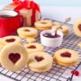 Biscuiți cu forme de inimioară și jeleu de zmeură pe un grătar, alături de o cană roșie, o cutie de cadouri și un bol cu dulceață