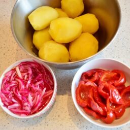 Cartofi fierți în bol de inox alături de un bol cu ceapă roșie marinată și un bol cu gogoșari murați
