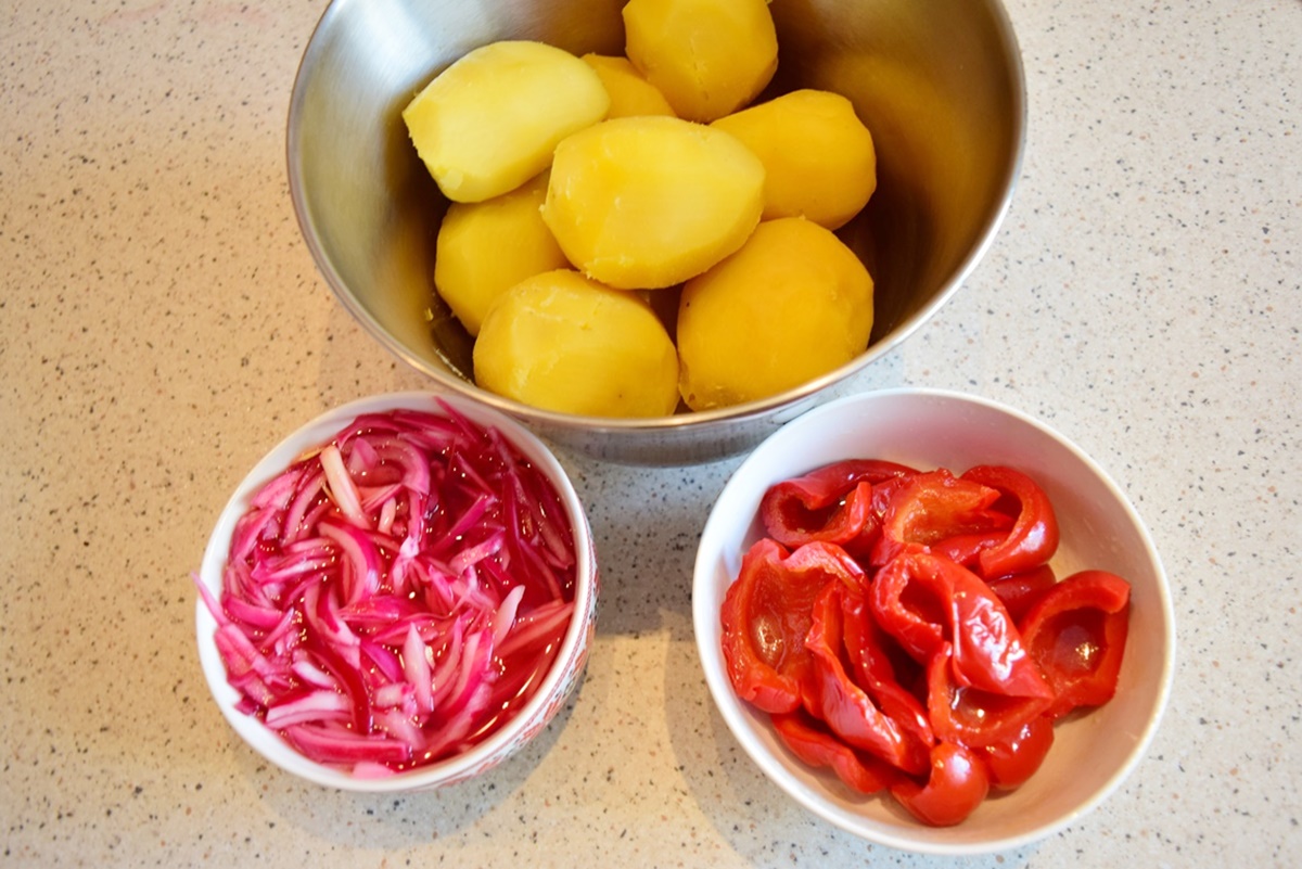 Cartofi fierți în bol de inox alături de un bol cu ceapă roșie marinată și un bol cu gogoșari murați
