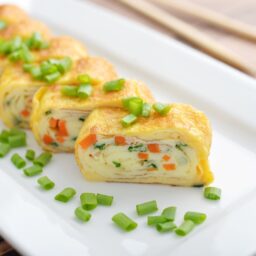 Ruladă din omletă cu morcovi și brânză porționată pe un platou alb și decorată cu rondele de ceapă verde
