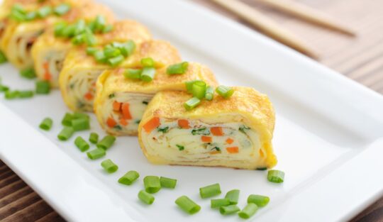 Ruladă din omletă cu morcovi și brânză porționată pe un platou alb și decorată cu rondele de ceapă verde