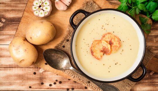 Supă cremă de usturoi cu cartofi și țelină decorată cu chipsuri de cartofi, în bol, alături de o lingură, doi cartofi și usturoi proaspăt
