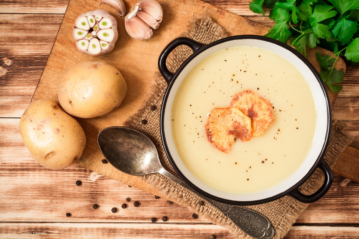 Supă cremă de usturoi cu cartofi și țelină decorată cu chipsuri de cartofi, în bol, alături de o lingură, doi cartofi și usturoi proaspăt
