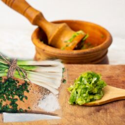 O legătură de usturoi verde alături de o lingură de lemn cu usturoi pisat, pe un tocător și un mojar cu pistil