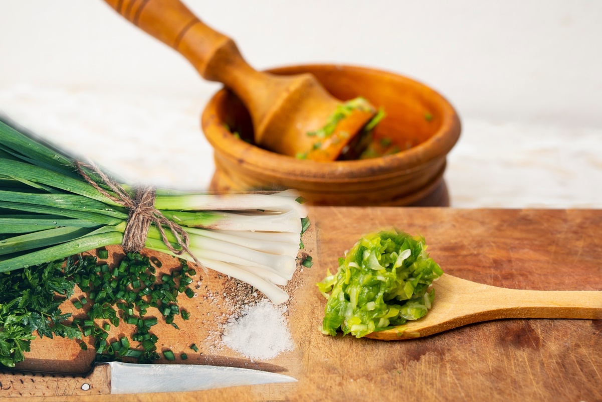 O legătură de usturoi verde alături de o lingură de lemn cu usturoi pisat, pe un tocător și un mojar cu pistil