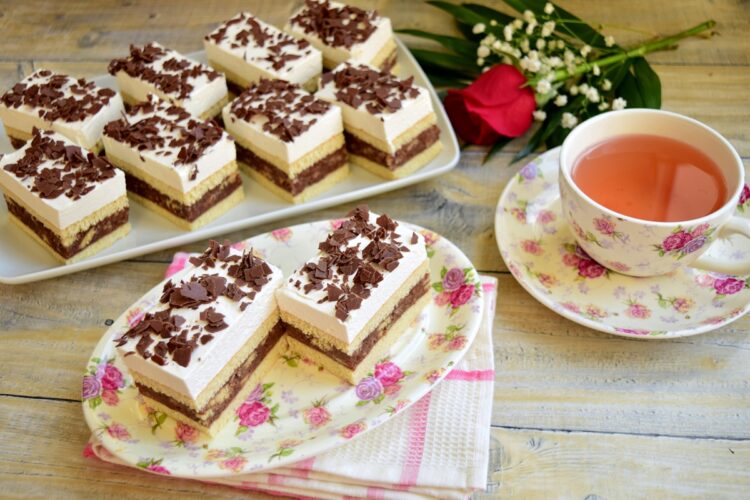 Prăjitura Kati porționată pe platouri alături de o ceașcă cu ceai și un buchet de flori