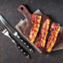 Trei felii de bacon puse pe un tocător de lemn alături de un cuțit și o furculiță