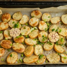 Rondele de cartofi cu o căpățână de usturoi tăiată în jumătăți și rondele de ceapă verde pe hârtie de copt