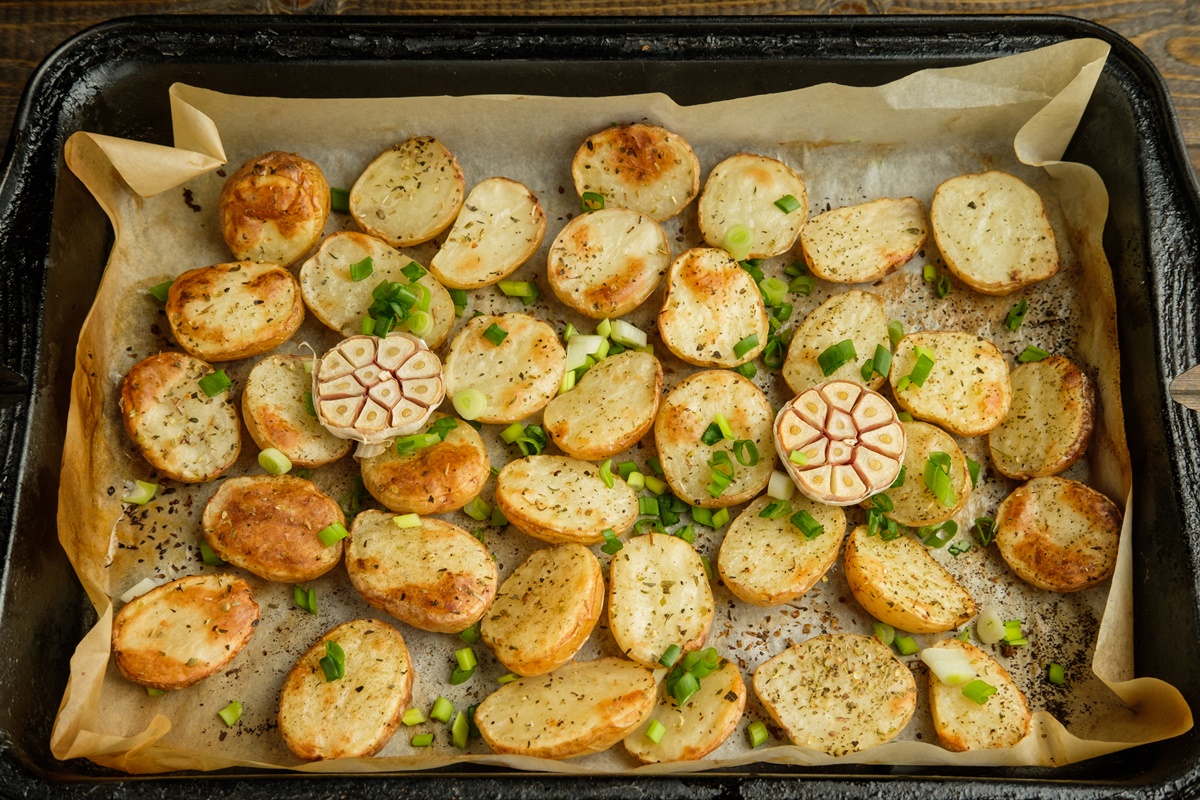Rondele de cartofi cu o căpățână de usturoi tăiată în jumătăți și rondele de ceapă verde pe hârtie de copt