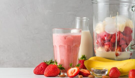 Pahar cu milkshake alături de o sticlă cu lapte, un recipient cu banane și căpșuni și fructe proaspete