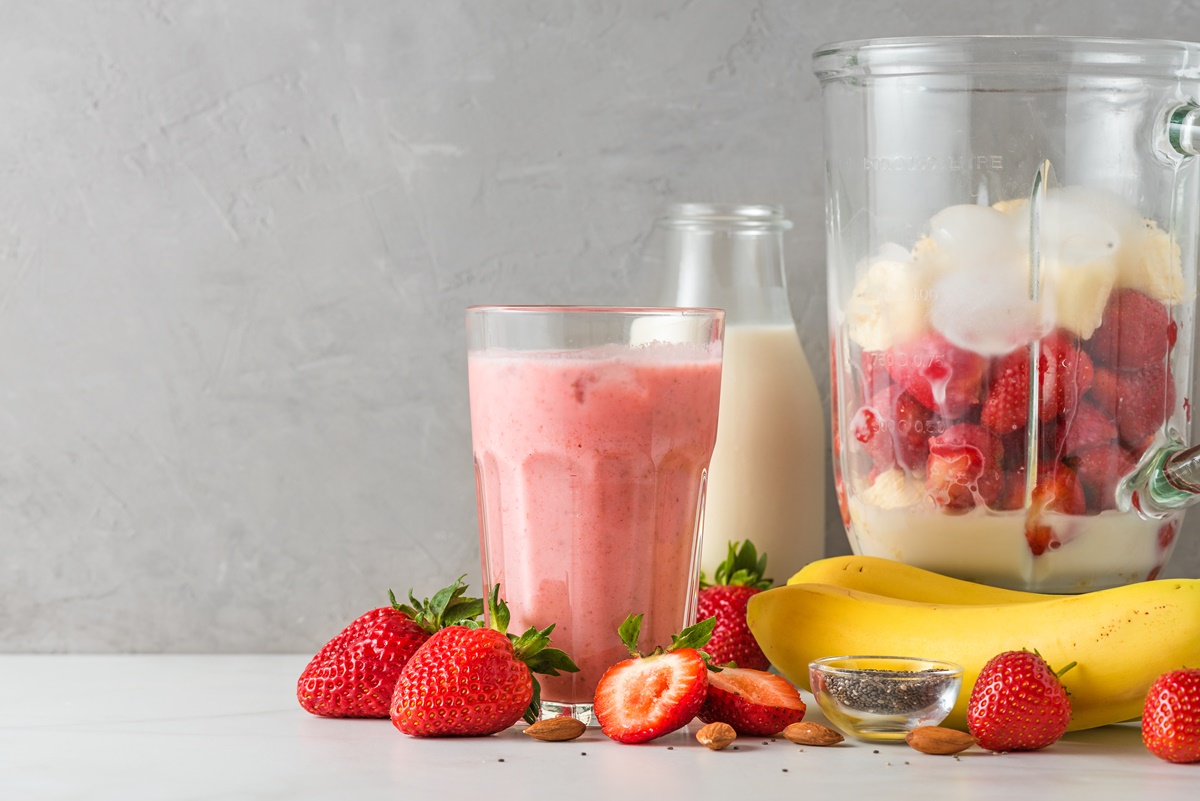 Pahar cu milkshake alături de o sticlă cu lapte, un recipient cu banane și căpșuni și fructe proaspete