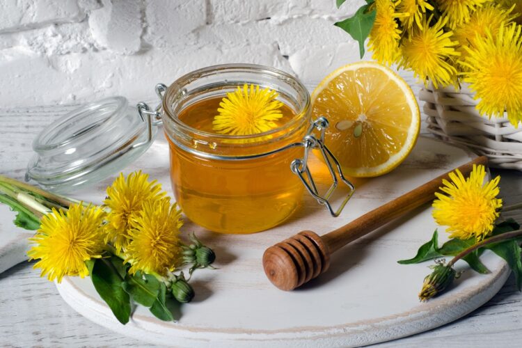 Sirop concentrat de păpădie denumit și miere falsă în borcan, alături de flori de păpădie și un tacâm de lemn pentru miere