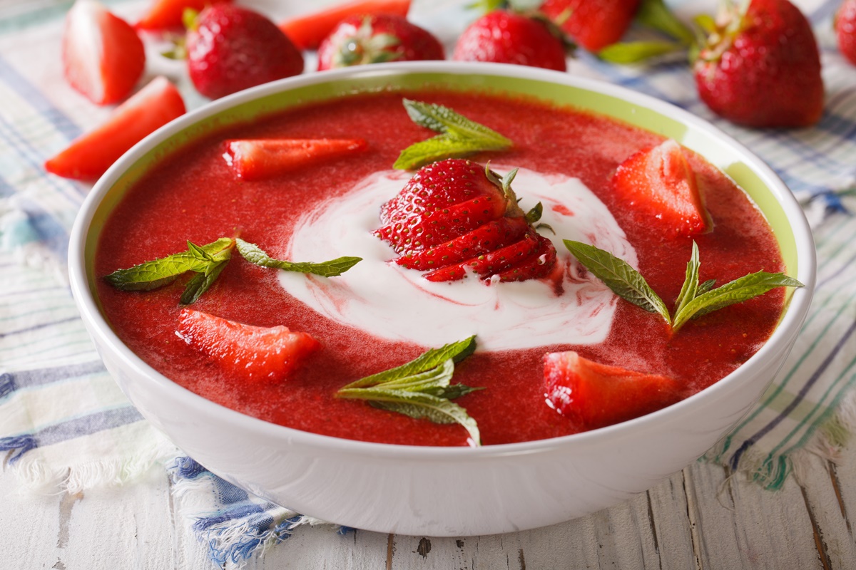 Bol alb cu supă rece de căpșuni cu mentă și smântână, decorată cu bucăți de căpșuni și frunze de mentă
