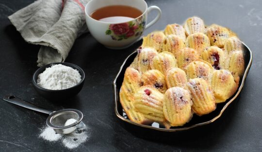 Madlene cu cireșe și vanilie pe un platou negru, alături de o ceașcă cu ceai, un bol cu zahăr și o sită