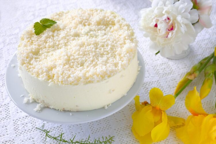 Cheesecake cu cocos pe un platou alb, o vază cu bujori și iriși galbeni