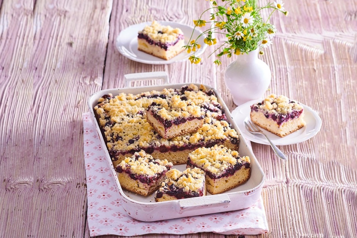 Prăjitură cu afine și crumble cu migdale porționată în tavă și pe două farfurii, alături de o vază cu flori