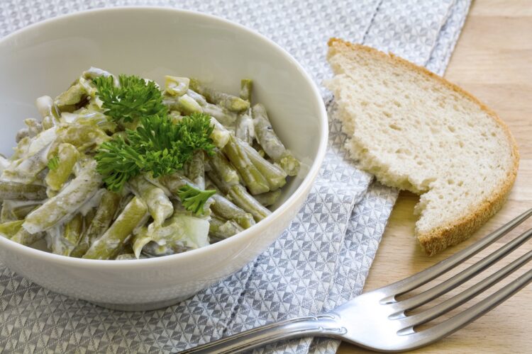 Salată de fasole verde cu iaurt grecesc și usturoi, decorată cu pătrunjel, în bol alb cu furculiță și o felie de pâine