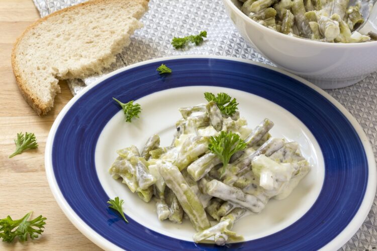 Salată de fasole verde cu iaurt grecesc și usturoi, decorată cu pătrunjel verde, în farfurie cu alb și albastru și în bol alb, alături de o felie de pâine
