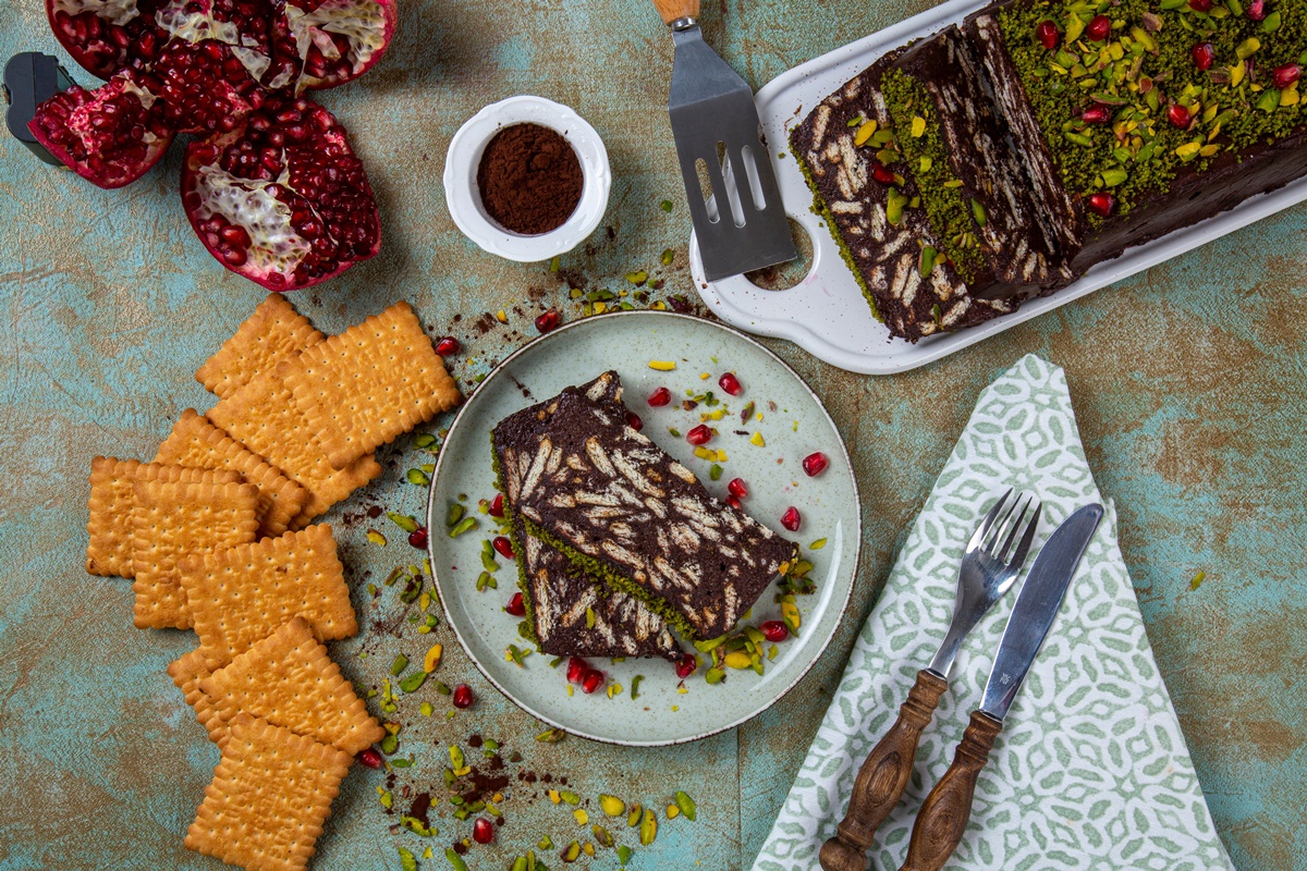 Tort de biscuiți cu glazură de ciocolată și fistic porționat pe platou și pe farfurie, alături de o rodie secționată, un bol cu pudră de cacao, biscuiți și o spatulă