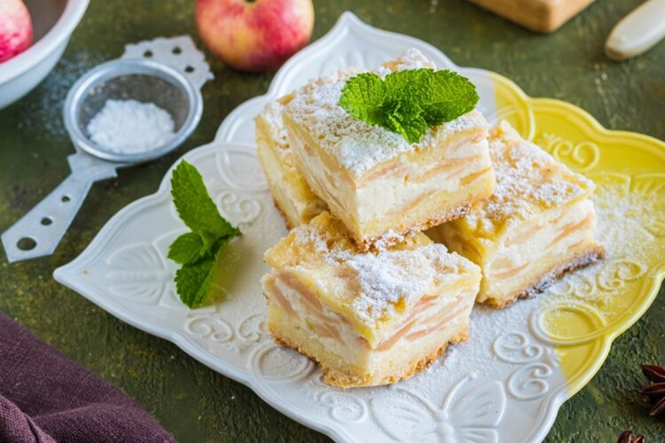 Prăjitură cu mere și brânză porționată pe o farfurie, alături de o sită cu zahăr și un măr