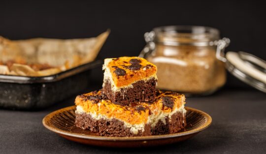 Porții de brownie cu brânză și dovleac pe farfurie, alături de tava cu prăjitură și un borcan cu zahăr
