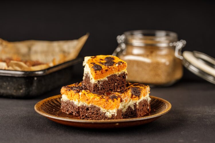 Porții de brownie cu brânză și dovleac pe farfurie, alături de tava cu prăjitură și un borcan cu zahăr