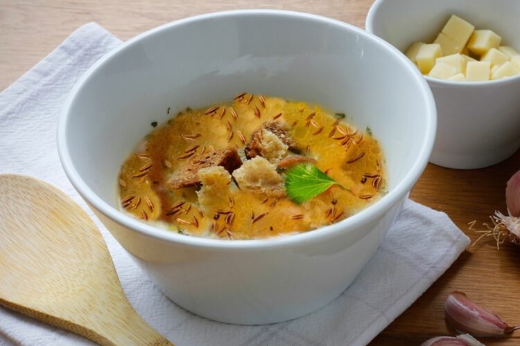 Bol cu supă de chimen și crutoane, alături de un bol cu cuburi de brânză și grăunți de usturoi