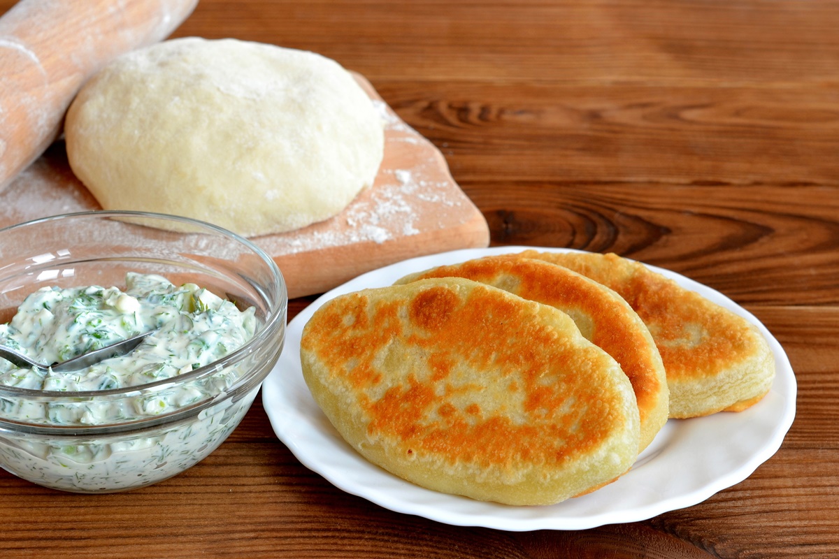 Trei plăcinte cu brânză și verdeață pe o farfurie, un bol cu sos de iaurt și aluat dospit pe un rocător