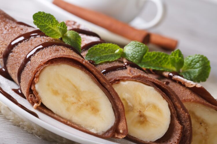 Clătite cu ciocolată și banane secționate pe o farfurie și decorate ci frunze de mentă