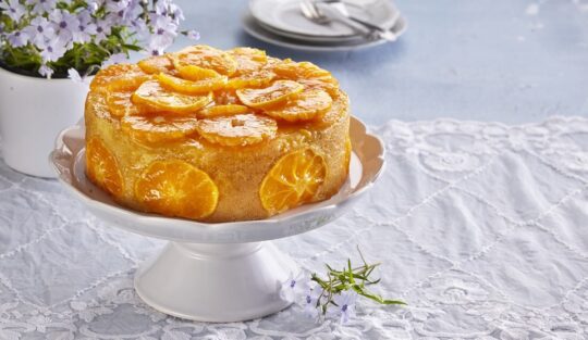 Tort cu mandarine pe un platou alb cu picior, alături de farfuriuțe cu tacâmuri și o vază cu flori