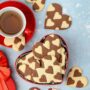 Biscuiți cu inimioare în două culori înt-o cutie de cadouri în formă de inimioară, alături de o ceașcă cu ceai
